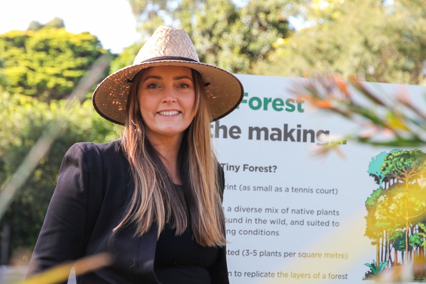 A young smiling woman wears a hat, dark top, stands in front of board saying forest in the making, greenery.