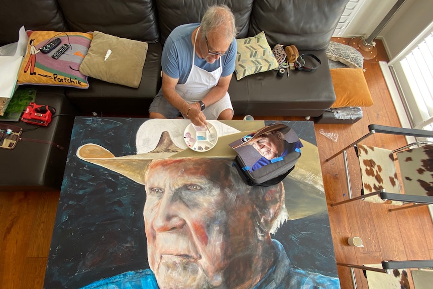 An aerial shot looking down at a man sitting on a couch painting a large portrait of an Indigenous man wearing an Akubra hat.