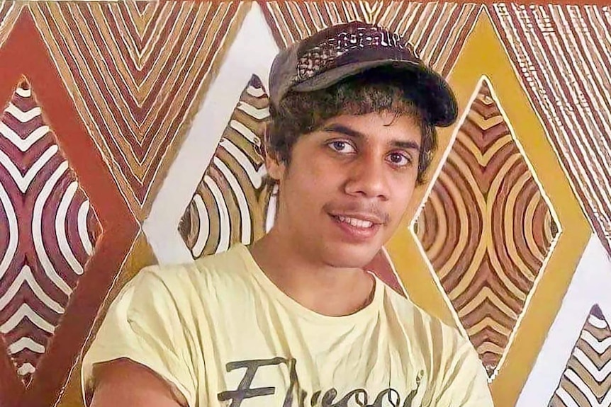 Man in yellow shirt with cap stands in front of Aboriginal painting.