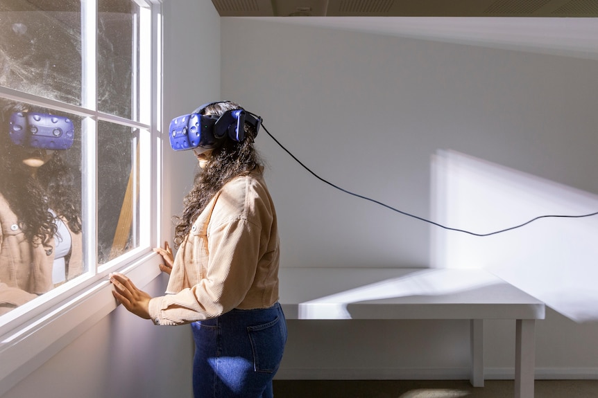 Woman with long dark curly hair wears taupe jacket, jeans and a blue virtual reality headset in white room in front of window.