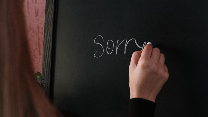 Woman wearing long-sleeved black top writing the word sorry on a chalkboard