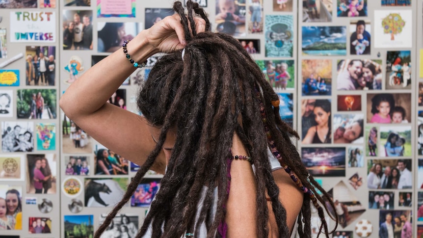 Keshira haLev Fife takes a hair band out of her dreadlocks, standing before photo wall.