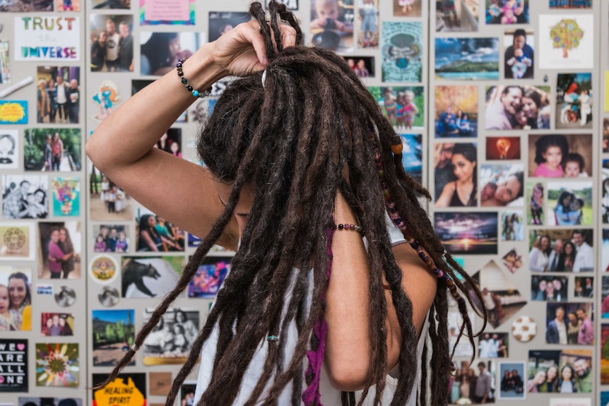 Keshira haLev Fife takes a hair band out of her dreadlocks, standing before photo wall.