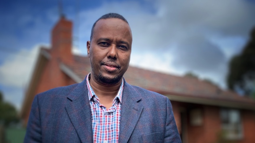 Ahmed Tohow stands in front of a suburban brick home, dressed in a jacket and chequered shirt.