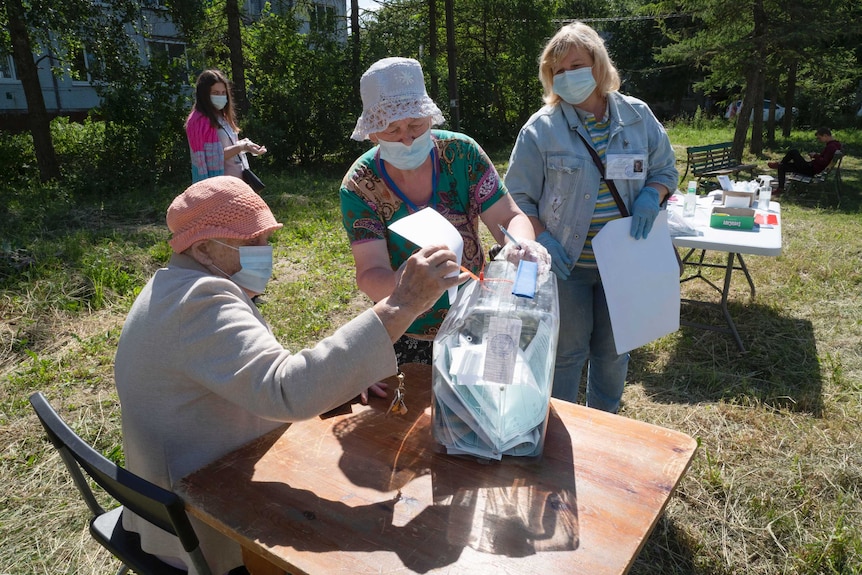 An old man wearing a mask and a hat puts a piece of paper in a clear box held by another older woman