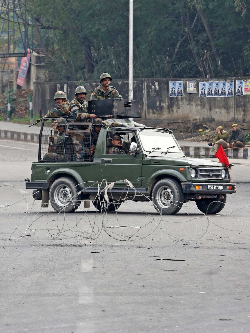 Indian Army soldiers in a vehicle patrol a street as a woman walks past.