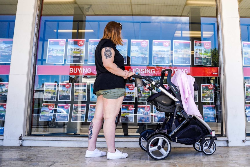 A woman stands with a pram looking at a large glass window with properties for sale pinned to it.