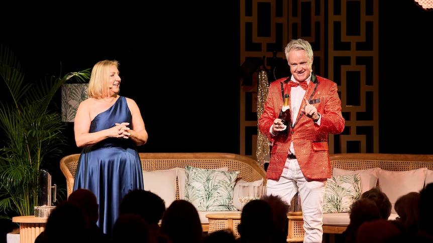 A woman with blonde hair wearing a blue dress, and a man in a red suit and bow-tie stand on a stage, smiling at the audience.