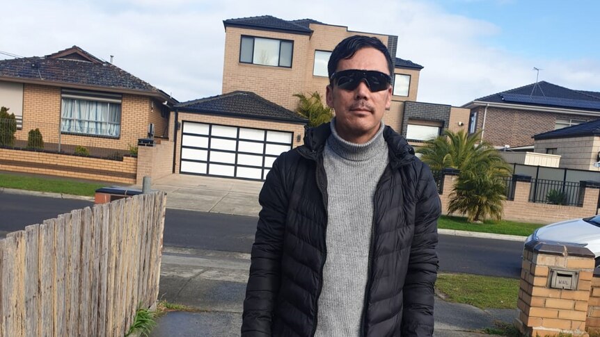 Man with black hair wearing black sunglasses, standing in a drive way, brick houses in the background