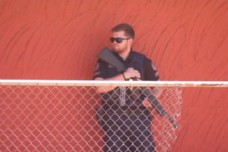 A police officer holding a rifle stands in the front of an orange wall and behind a fence