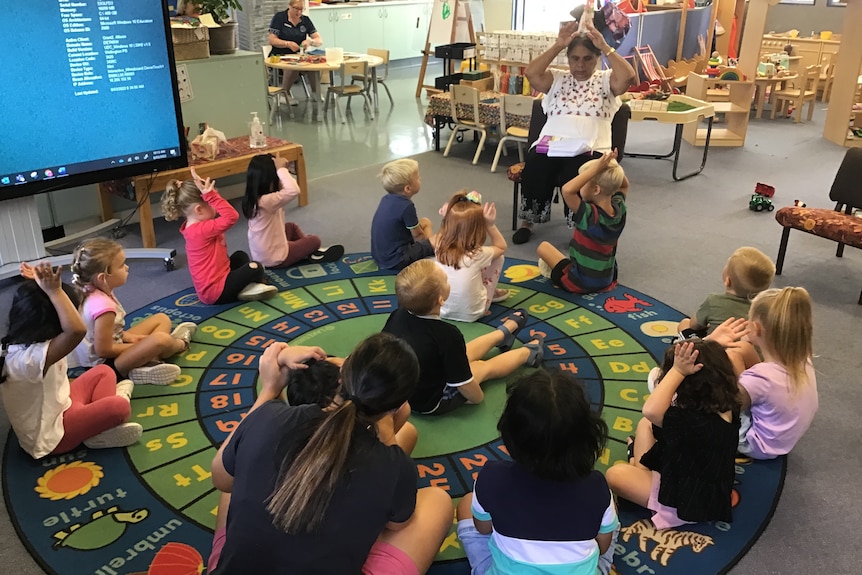 Children are sitting on a rug listening to their indigenous teacher. 
