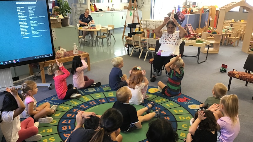 Children are sitting on a rug listening to their indigenous teacher. 