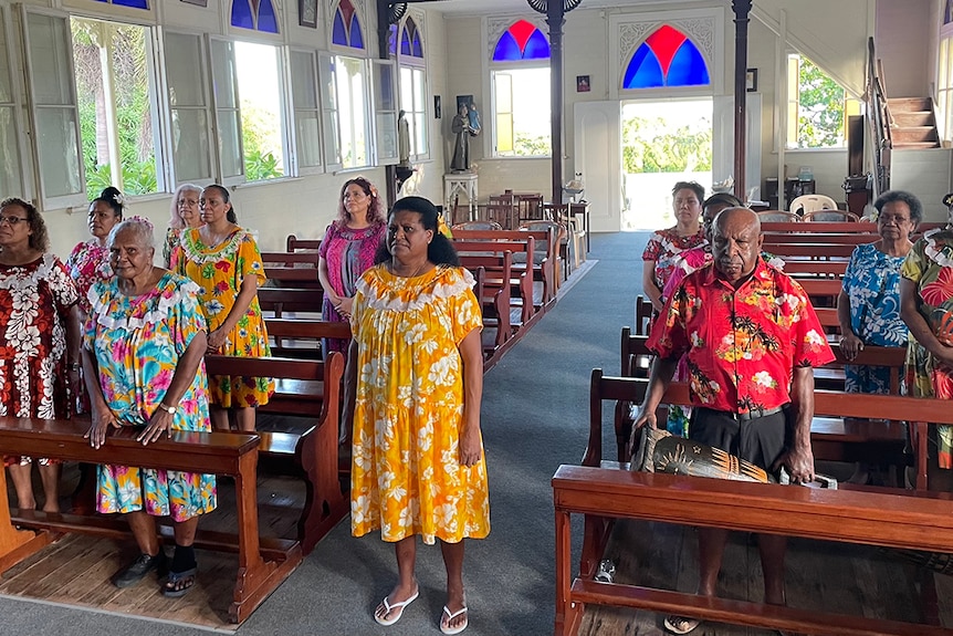11 women and one man stand in church pews wearing colourful clothing