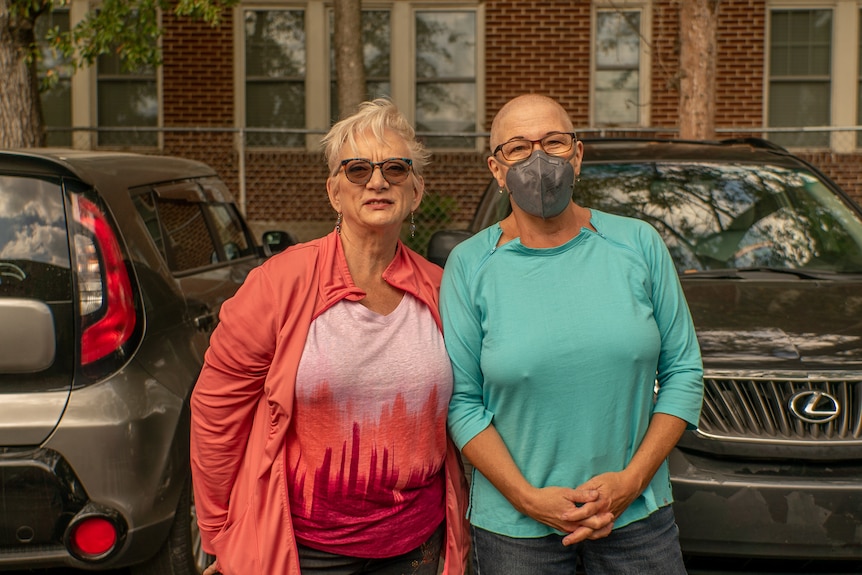 A woman in a blue shirt with a shaved head and grey Covid mask stands next to her wife who is wearing a red jacket and t-shirt