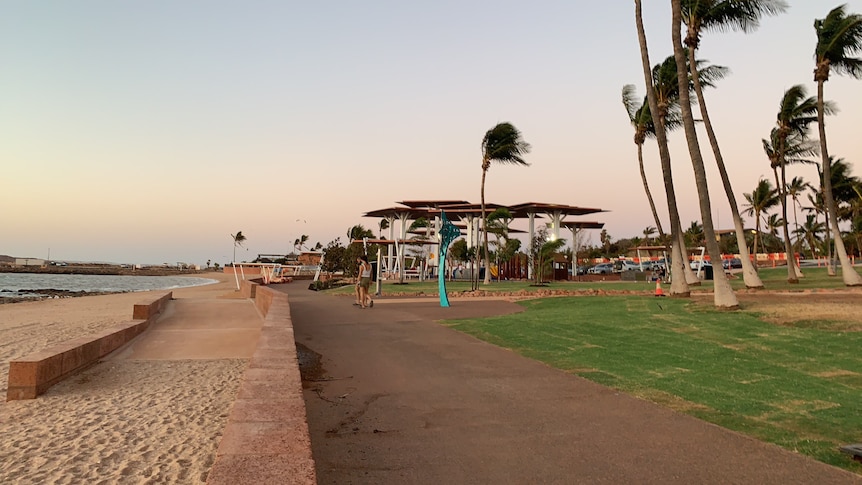 Sandy beach foreshore in early morning. Wind-blown palm trees, empty children's playground & two people on boardwalk in distance