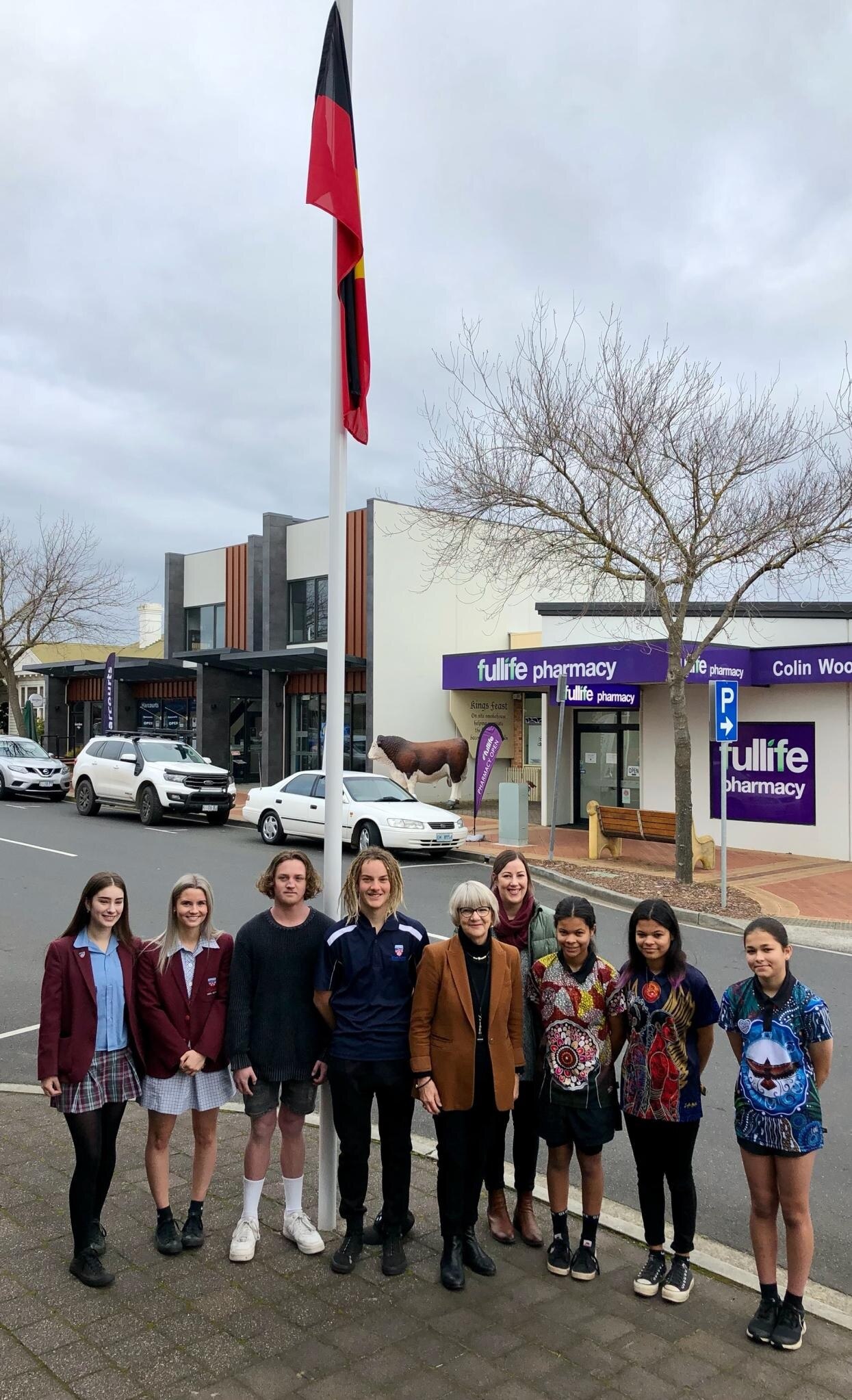 A group of students poses with female mayor at flag pole flying Aboriginal flag.