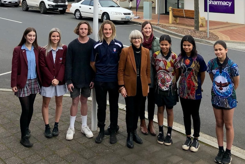 A group of students poses with female mayor at flag pole flying Aboriginal flag.