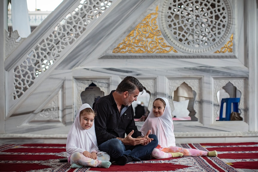 A father and his two daughters, both wearing hijabs, sit inside a mosque.