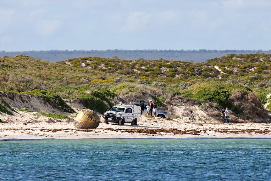 A long distance image of reporters and police standing near a piece of space junk washed up on a beach.