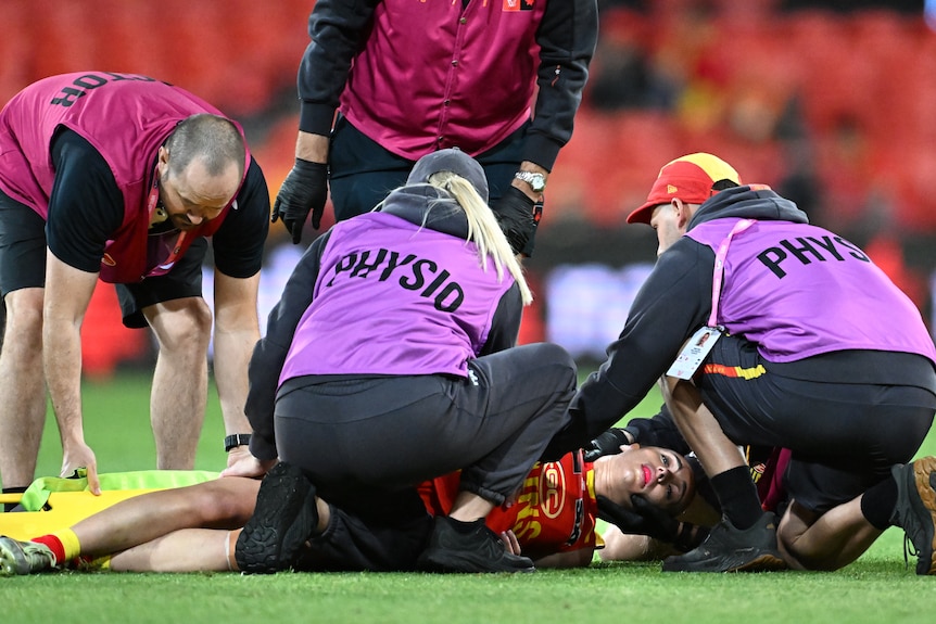 Four people wearing pink and purple shirts hunched over a woman lying on a green grass field being dragged onto a stretcher