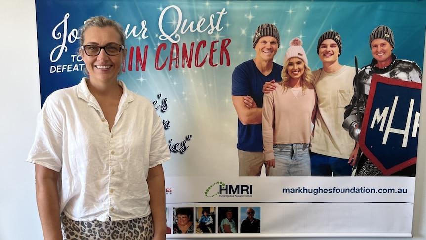 a woman in a white shirt smiling in front of a Mark Hughes Foundation poster