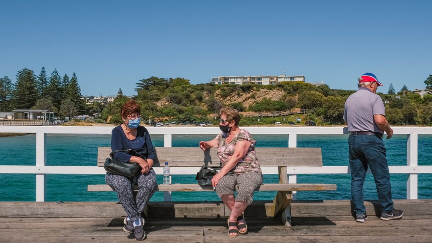 Two people wearing face masks sit and talk on a bench on a sunny day with the blue sky and blue ocean behind them.
