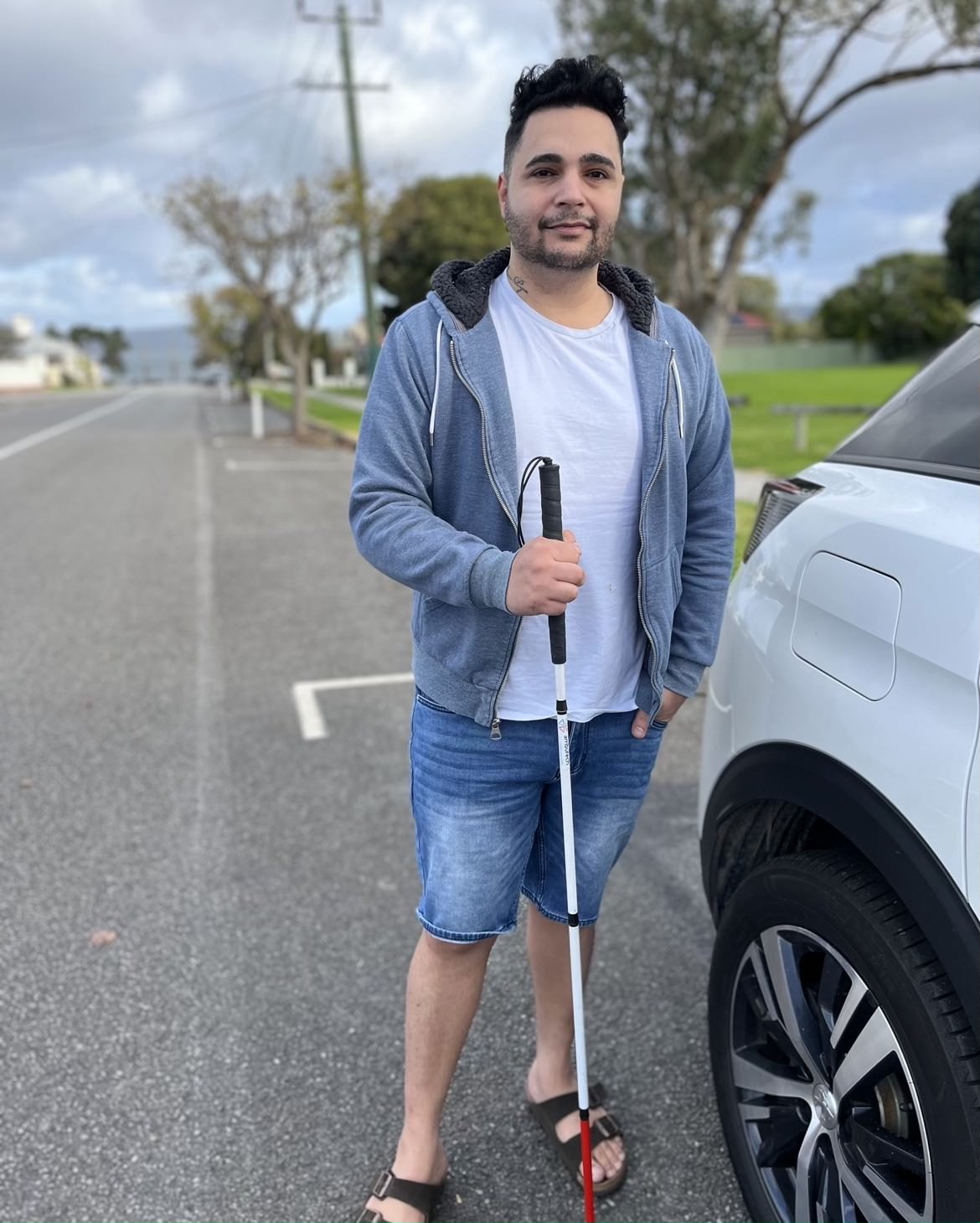 Man with vision assistance cane stands beside a car with beach in the background.