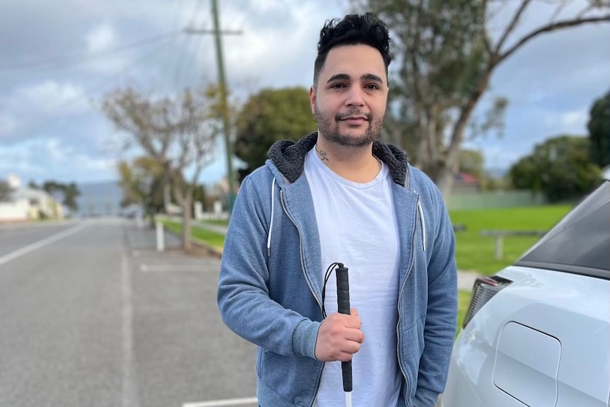 Man with vision assistance cane stands beside a car with beach in the background.