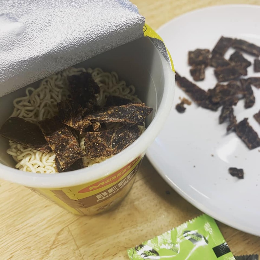 A cup of dry instant noodles with beef jerky in it, waiting for water to be added.