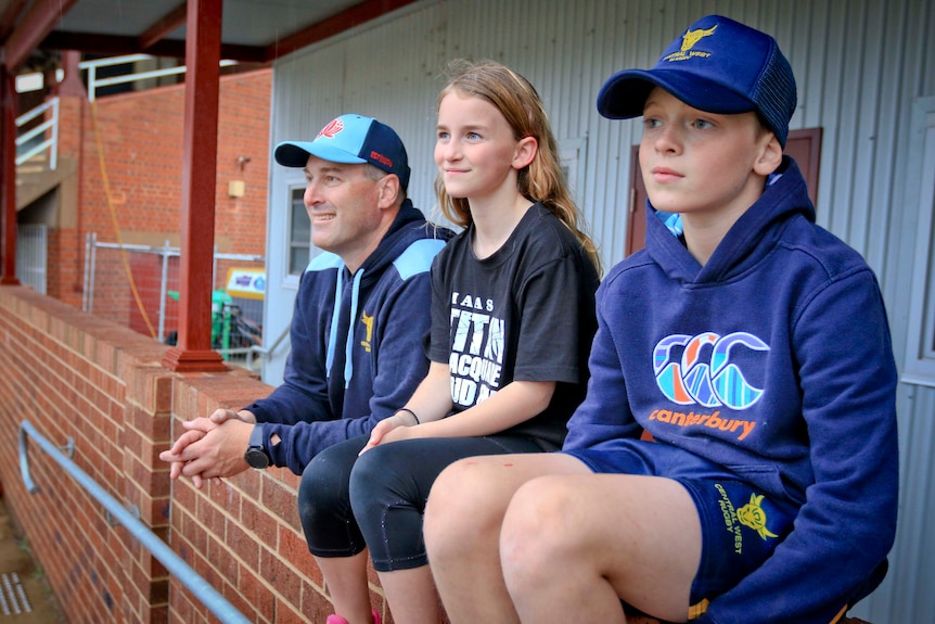 Justin and his two school-aged kids on the sidelies of a footy oval, watching the action.