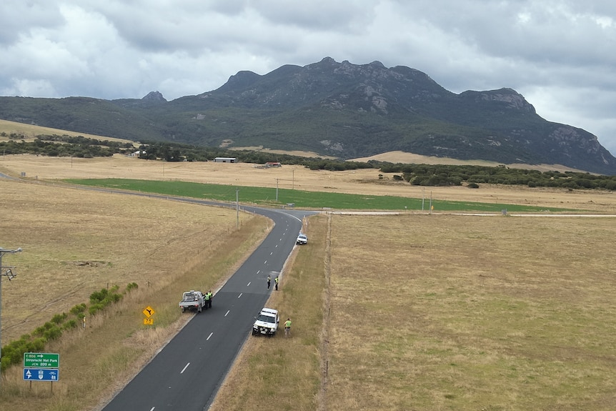 Three police vehicles are seen on the side of a road. A large mountain looms in the distance.