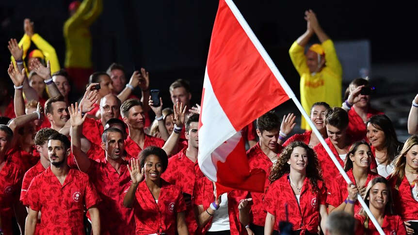 A team of people dressed in red and white walk and wave through a stadium, with a Canadian flag.