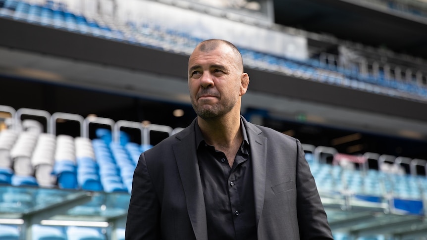 A man in a black suit and shirt stands in front of a football stadium of seating 