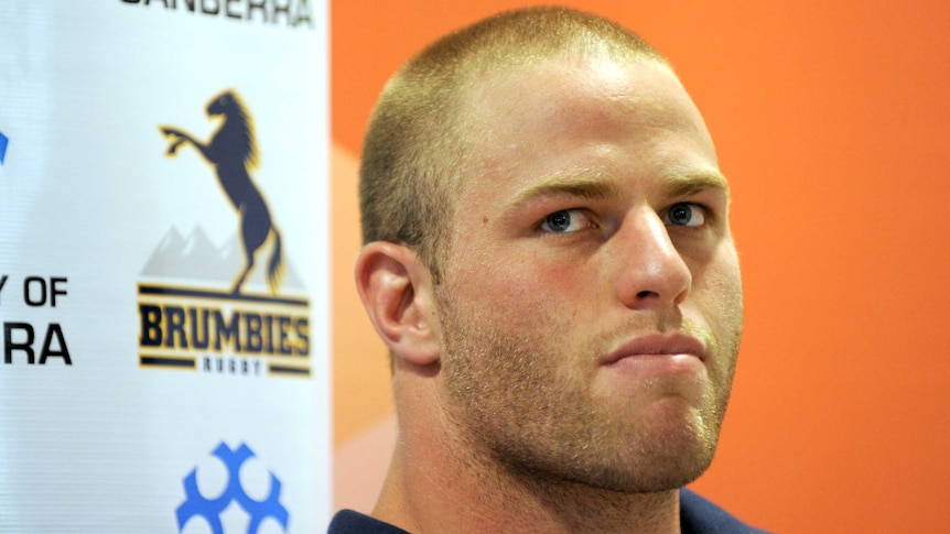 A rugby union player sits at a press conference in front of a Brumbies sponsors' banner.