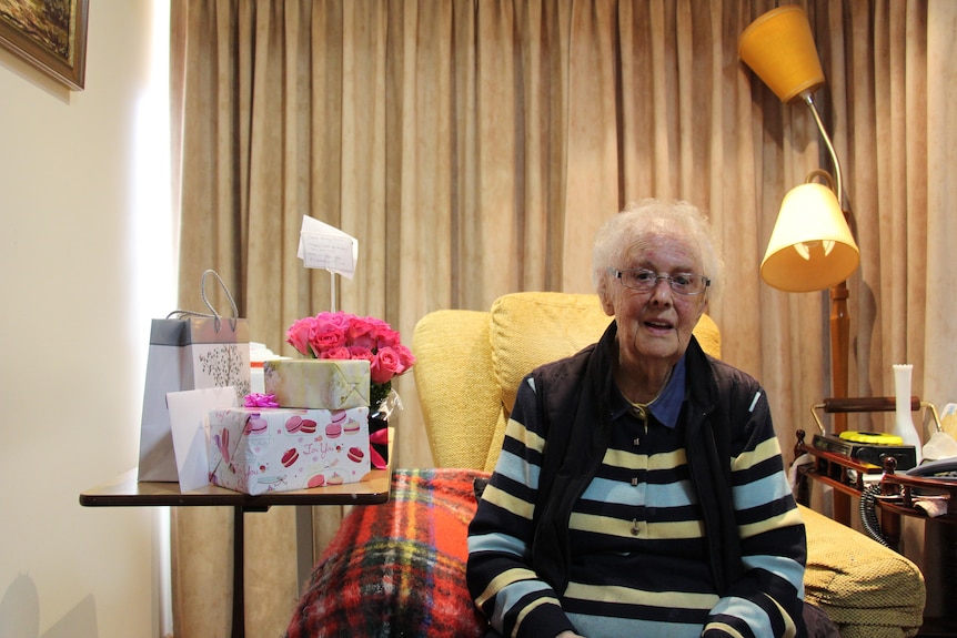 A woman with white hair in a striped jumper looks at the camera, sitting in an armchair with presents and a lamp beside her