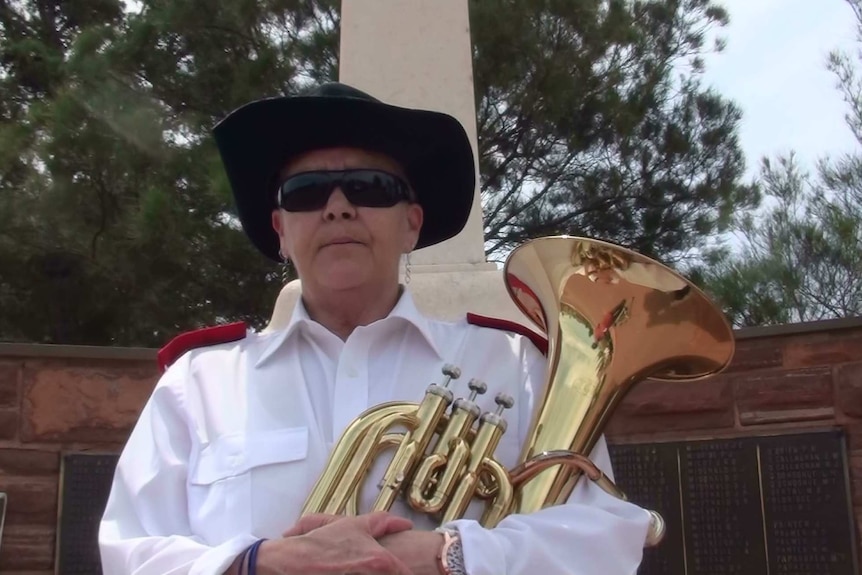 A woman wearing a white long-sleeve shirt, black sunglasses and a black hat holds a baritone horn.