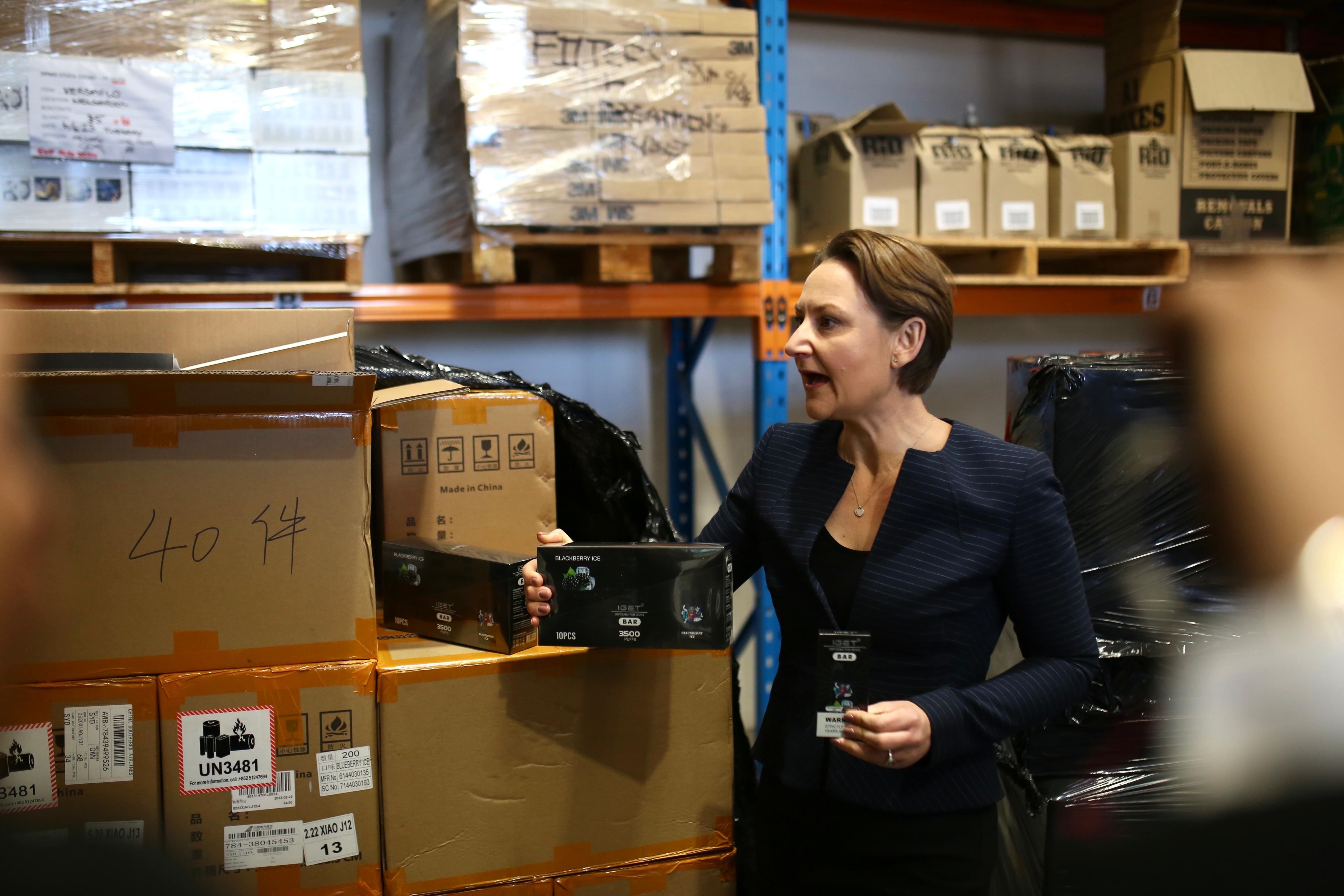 一名中等长度头发的女士在仓库里拿着一盒电子烟。 她穿着一件海军蓝西装外套。