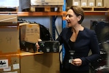 A woman with medium length hair holds a box of vapes in a warehouse. She is wearing a navy blazer.