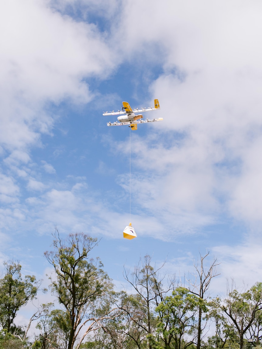 a white drone in the air with a package attached underneath it