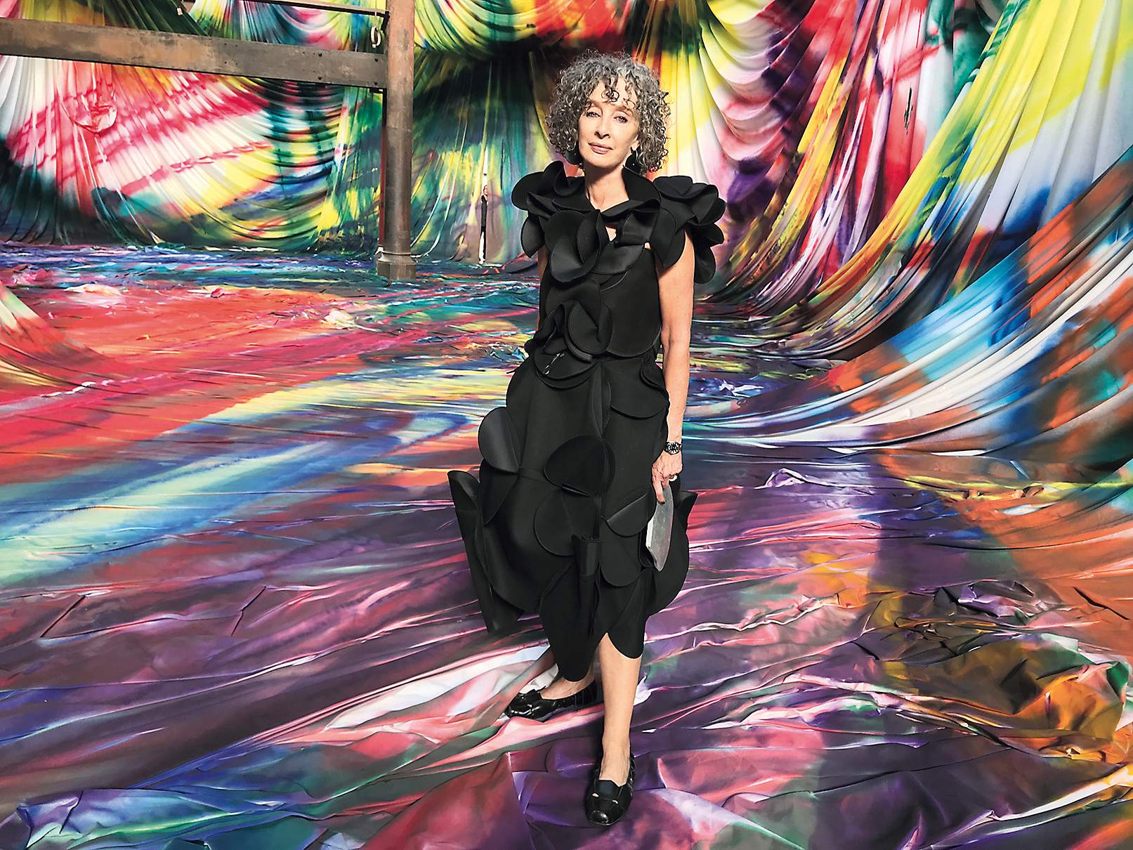 Anna Schwartz, a middle-aged woman with grey curly hair wearing a black dress in front of a colourful textile artwork