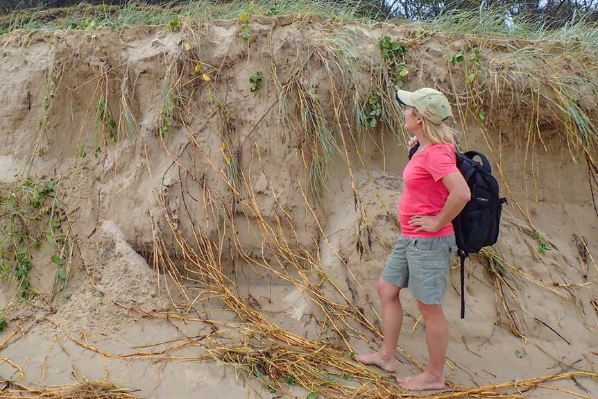 A woman stands in front of an eroded sand dune on a beach.