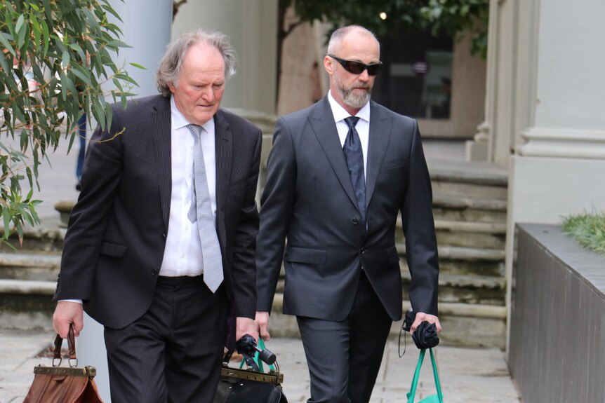Two balding men wearing suits and ties and carrying bags walk walk side by side outside a building.