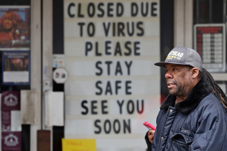A man in the foreground with a cap and dreadlocks with a building in the background with sign reading 'closed due to virus'