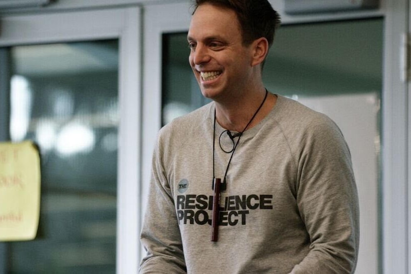 A man in a jumper with text "The Resilience Project" smiles while standing