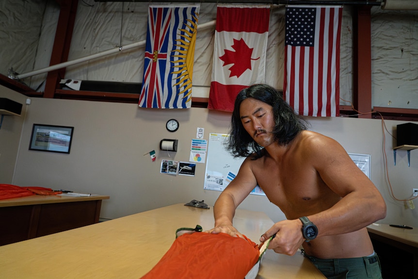 A shirtless man works tying parachute fabric on a workbench. The British Columbia, Canadian and US flags hang behind him.