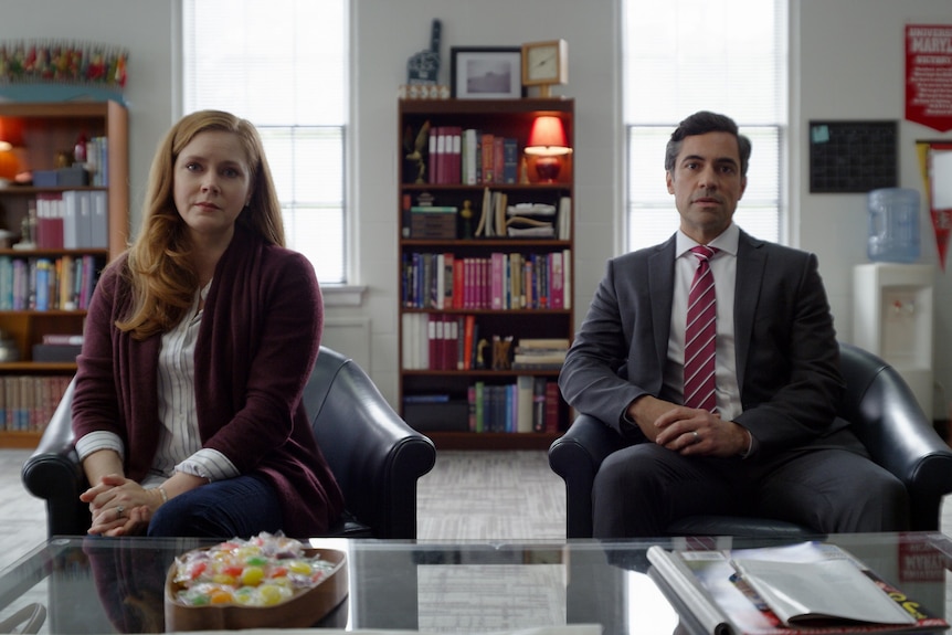 A couple in their 40s sit looking sombre with their hands folded in front of them in a principal's office