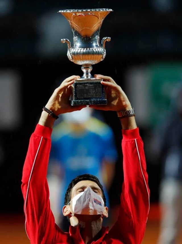 Novak Djokovic holds a trophy aloft while wearing a face mask.