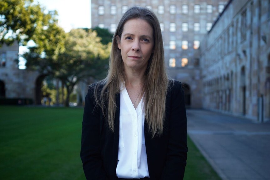 A woman wearing a white shirt and black blazer stands in the grounds of UQ