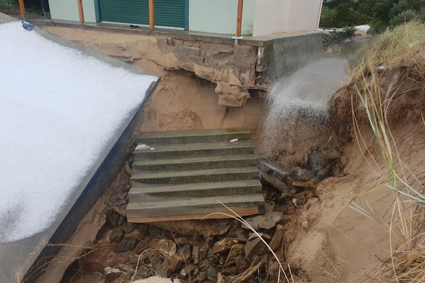 A damaged building on a beach with its foundations showing and displaced concrete stairs.