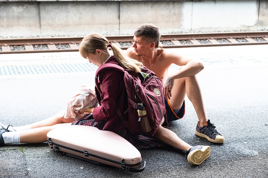 A ponytailed girl in maroon school blazer, backpack and dress sits on ground of train platform with topless boy with rat tail.